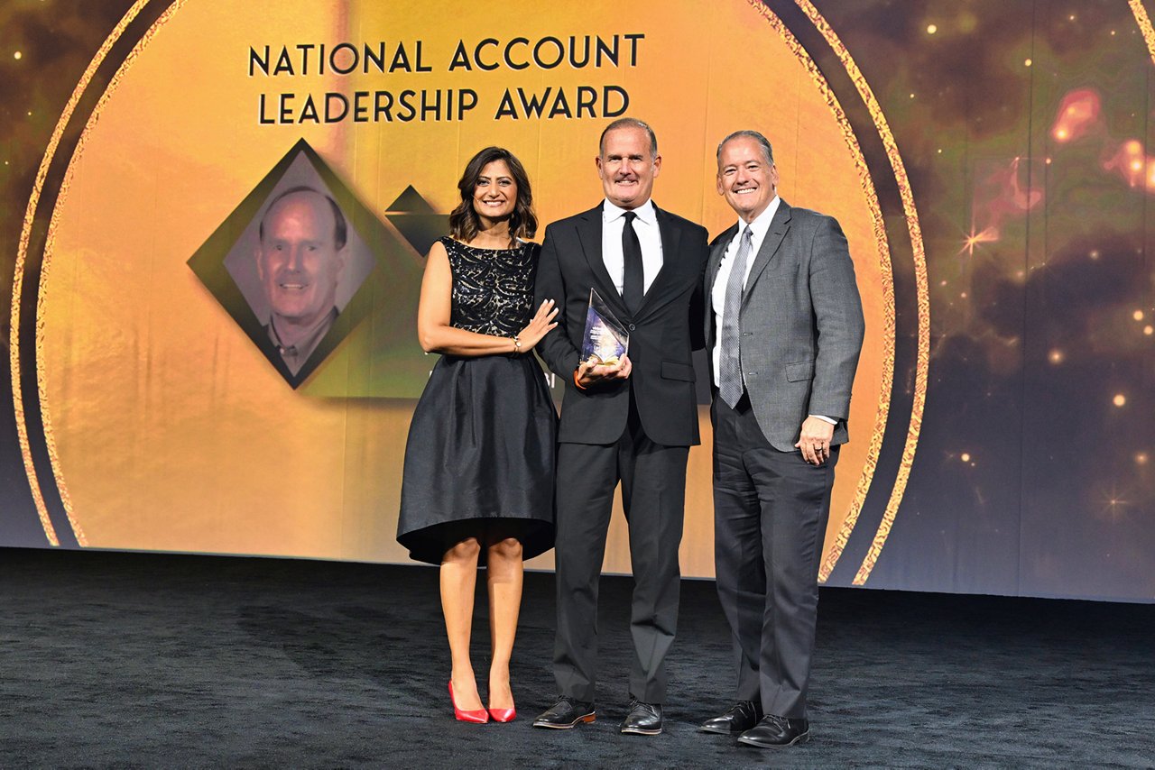 Award ceremony with three individuals on stage in front of a “National Account Leadership Award” backdrop; central figure holds an award, with a portrait displayed in the background.