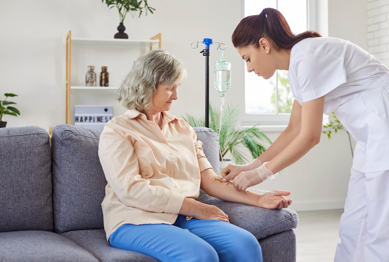 Senior patient getting intravenous vitamin therapy procedures at home. Professional geriatric nurse practitioner physician inserts catheter needle in vein of senior retired pensioner woman on sofa