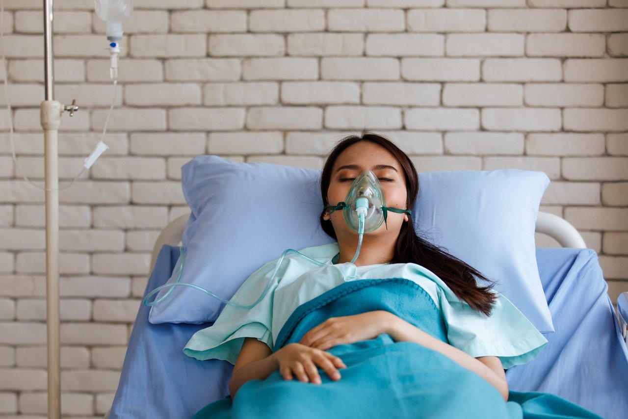 Young long black hair sick asian female patient wear green shirt and oxygen intubation mask on face sleeping on blue sheet hospital pillow and bed in icu ward room with white brick wall background.