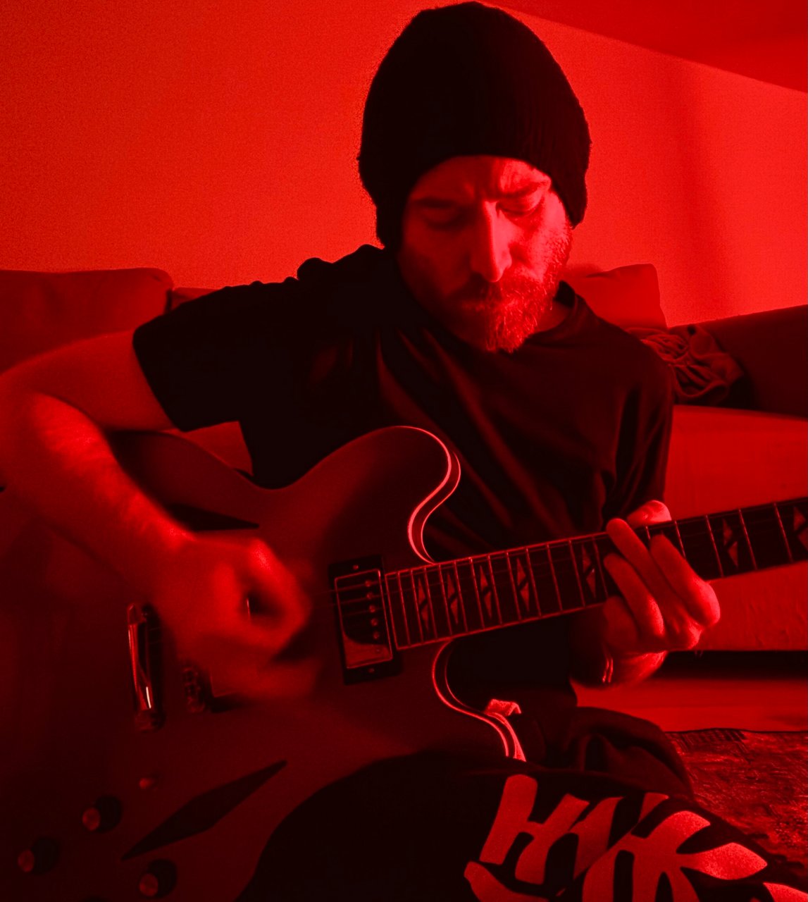 Andrea wears a beanie and plays guitar in red room lighting.
