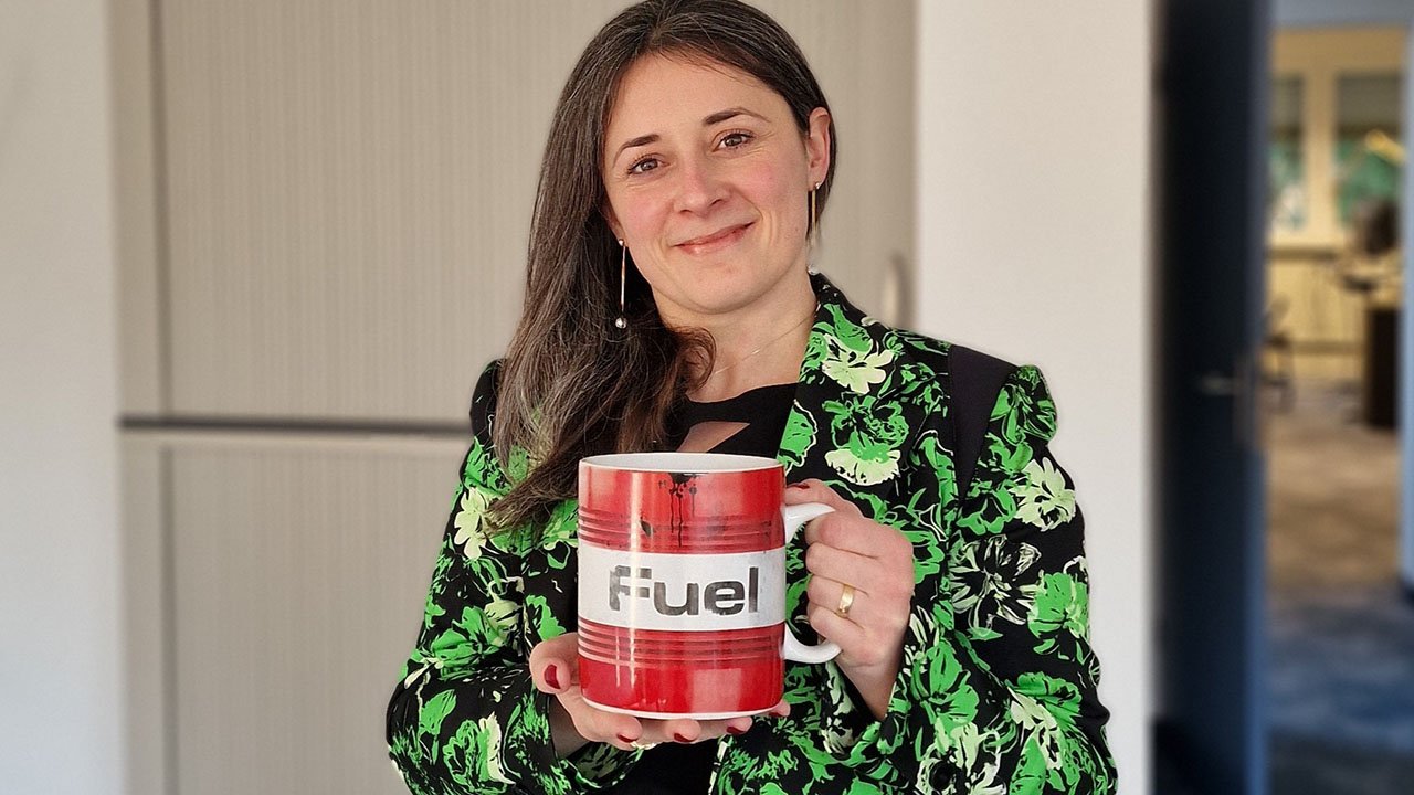 Beata Jonik-Nowak wearing a black blazer with a green floral pattern, smiling at the camera while holding a large red and white mug labeled 'Fuel'."