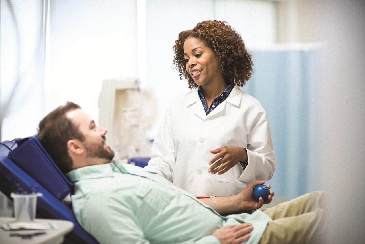 A man seated in a blood donor chair is talking with a nurse while squeezing a Fresenius Kabi foam ball and donating blood.