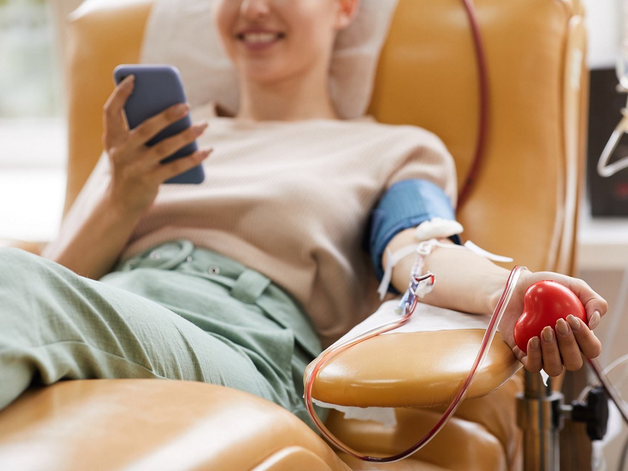 Close-up of a person donating blood while seated in a medical chair, holding a red stress ball. in one hand and her smartphone in her other hand.