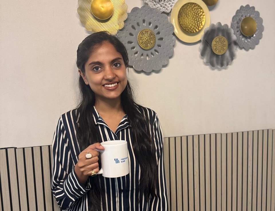 Nitya Shah wearing a striped shirt holding a Fresenius Kabi mug, standing in front of a decorative wall.