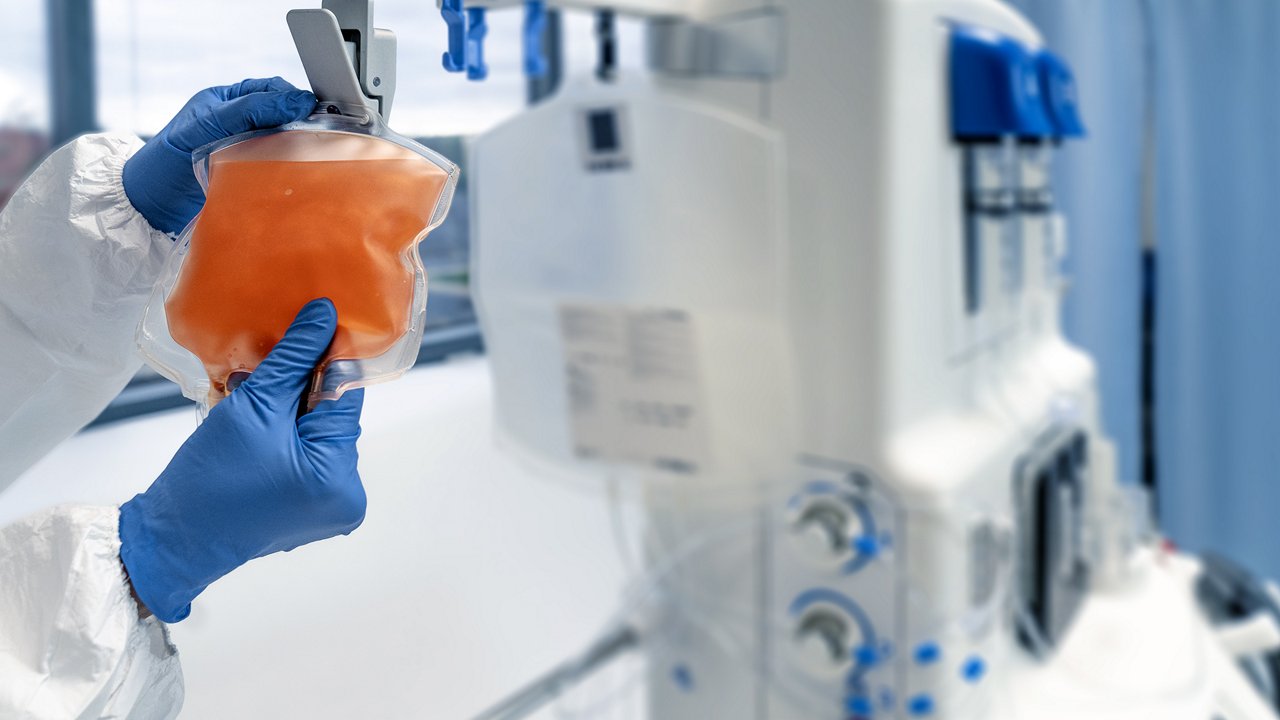 Lab technician in protective gear holds orange liquid-filled bag next to medical equipment in clinic.