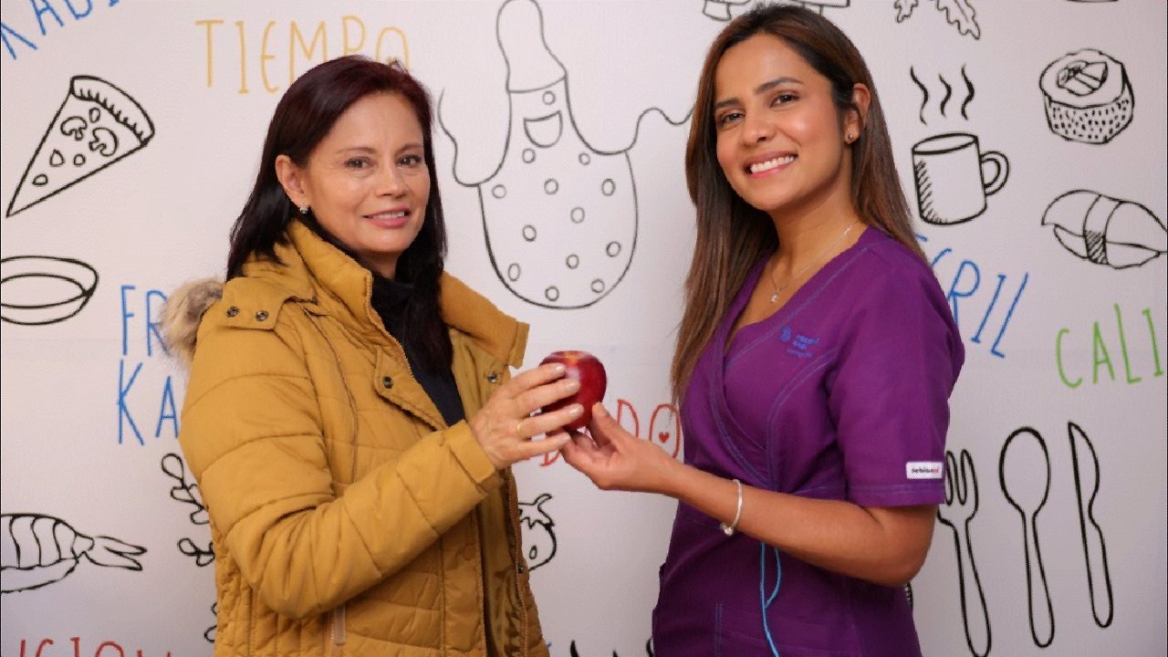 Diana and her dietician holding an apple in front of a food-themed wall.