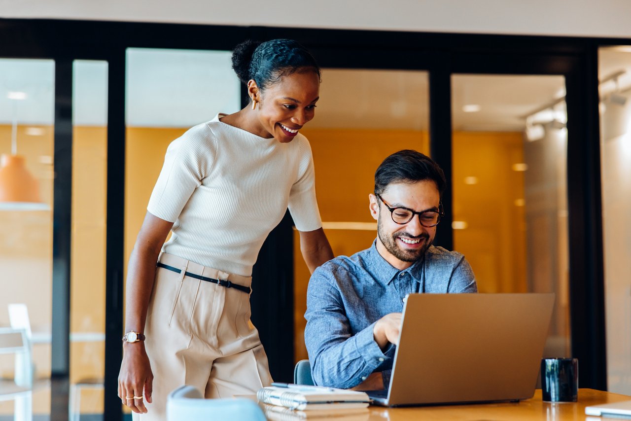 A cheerful man and woman work together on a laptop in a professional office setting. They appear to be discussing a shared project, fostering teamwork while enjoying their workplace environment.