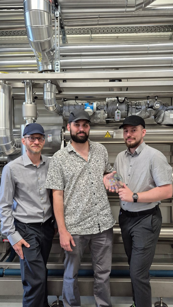 Three employees stand in front of stainless-steel industrial equipment, with one person holding a transparent Champion Supply 2026 award.