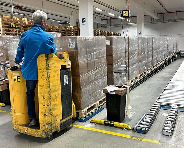 Worker in a blue jacket operates a yellow electric pallet jack, moving wrapped pallets in a warehouse with high shelves and safety markings.