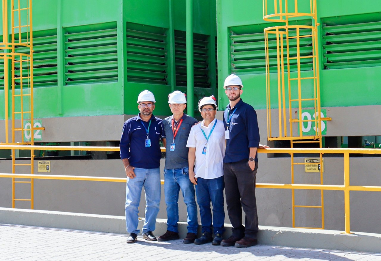 Four people wearing hard hats stand in front of large green industrial equipment with yellow safety ladders and railings.