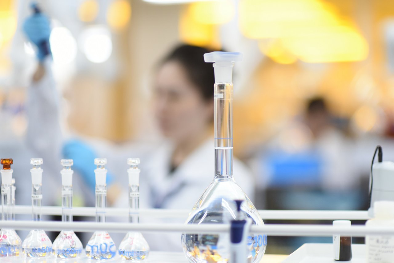 Laboratory workbench with volumetric flasks and test tubes, person in lab coat working in the background