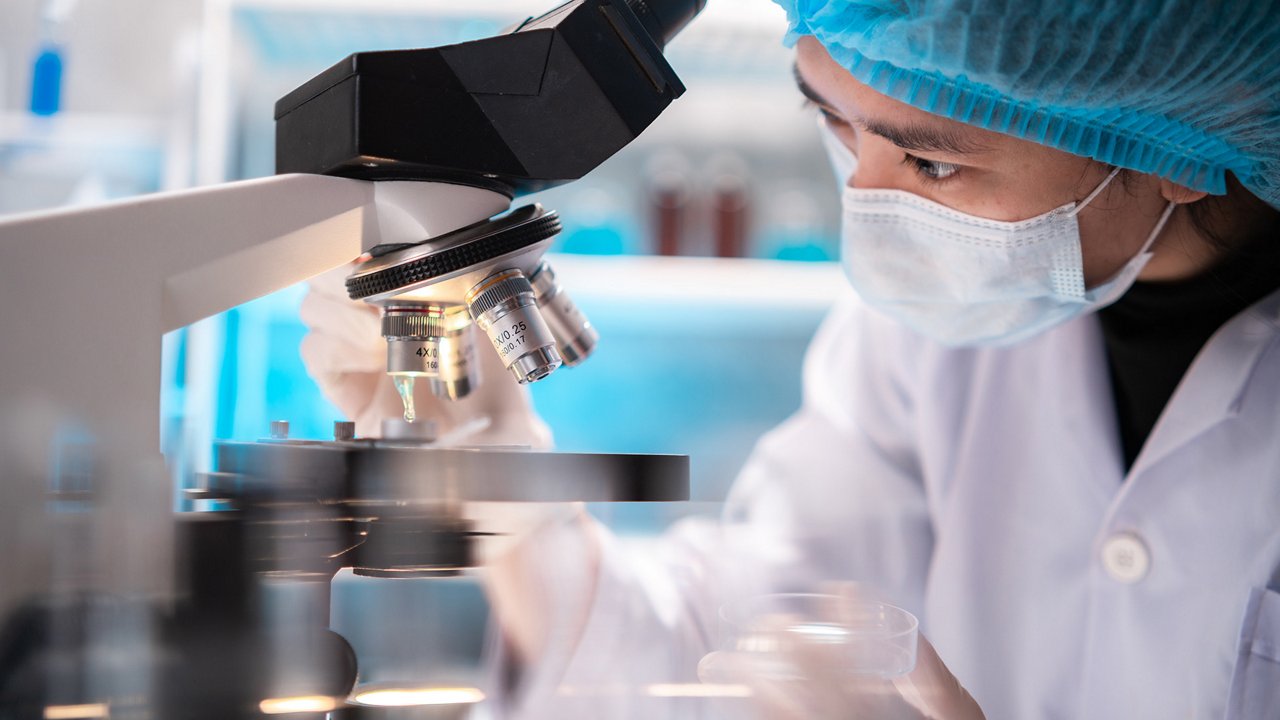A researcher in a lab coat and blue hair cover operates a microscope in a professional laboratory setting. The individual is adjusting the sample under observation, with various lab equipment and containers visible in the background, indicating an active research environment.