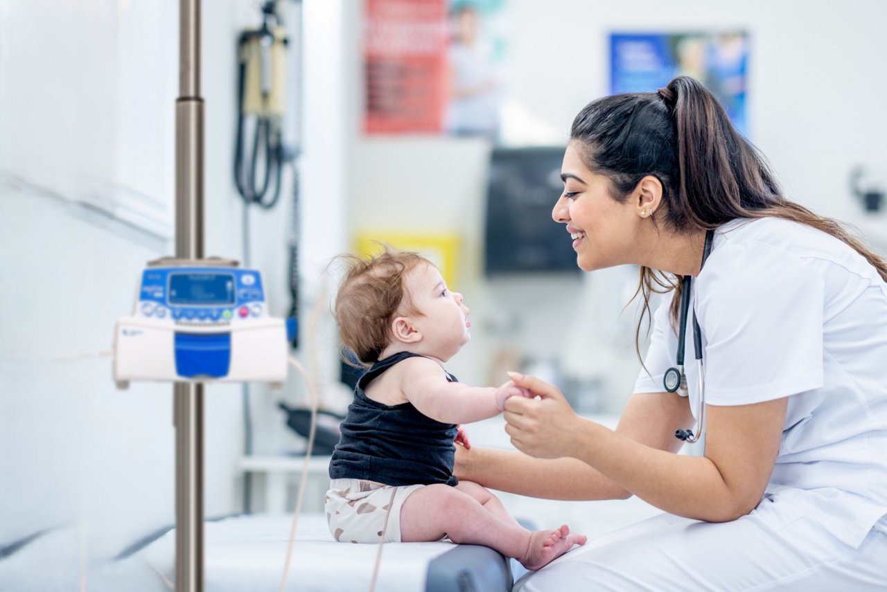 Healthcare professional wearing a white uniform and a stethoscope gently holds the hand of a sitting baby on an examination table in a clinical setting, with medical equipment and monitors visible in the background.