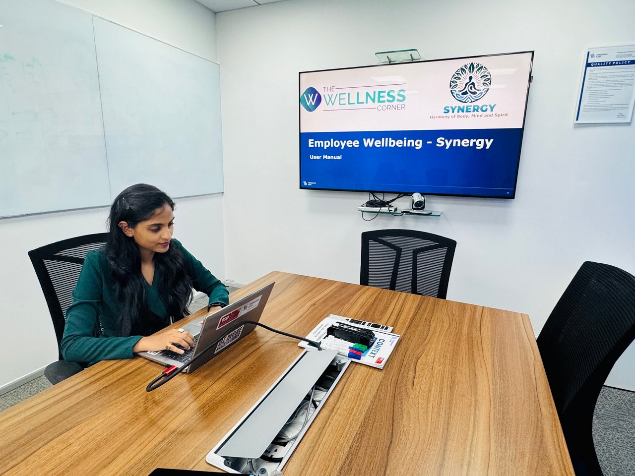 Nitya Shah working on a laptop in a meeting room, with a presentation on employee wellbeing shown on the screen.