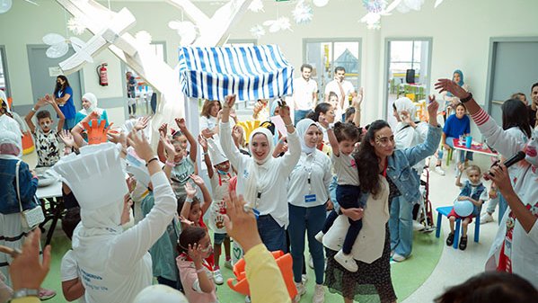 Group of people, including children and adults, gathered in a brightly lit room with decorations and costumes.