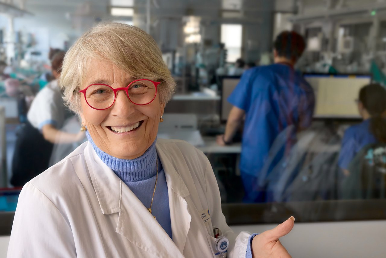 Prof. Luciana Teofili smiling, dressed in a white lab coat and blue turtleneck sweater, standing in front of a glass partition with a visible clinical or laboratory environment in the background, where healthcare personnel are working at computer stations