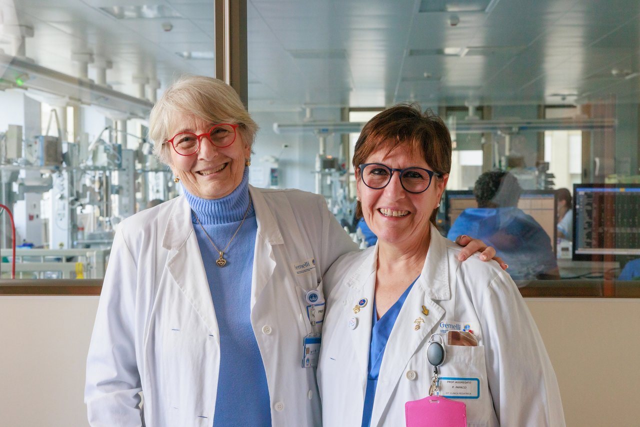 Prof. Luciana Teofili and Prof. Patrizia Papacci in white lab coats standing in front of a glass wall with a clinical setting and staff visible in the background.
