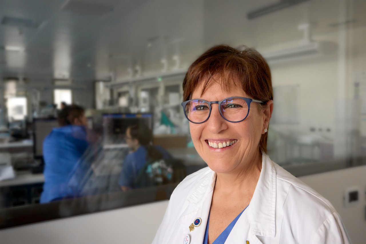 Prof. Patrizia Papacci dressed in a white lab coat and blue scrubs, standing in front of a glass partition with a view into a clinical or laboratory setting, where medical staff are working at computer stations in the background.