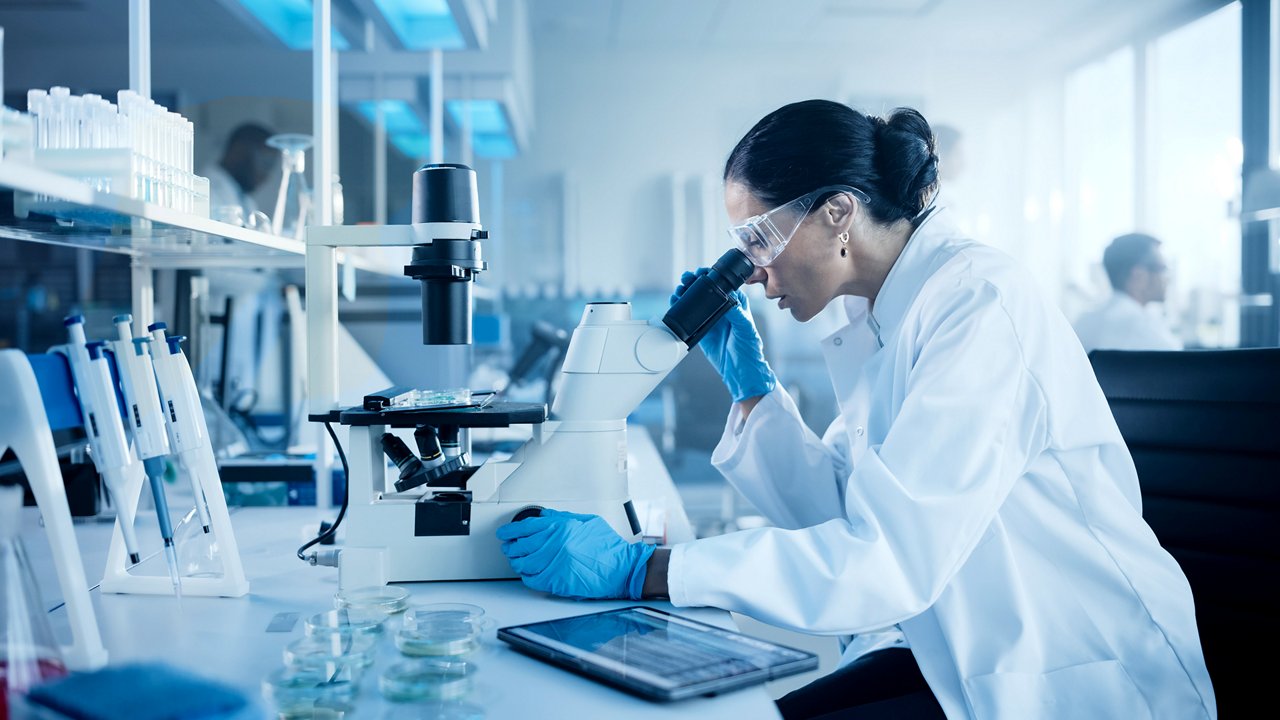 A scientist wearing a white lab coat and blue gloves examines a sample through a microscope in a modern laboratory. Several petri dishes and a tablet are on the table in front of her. 