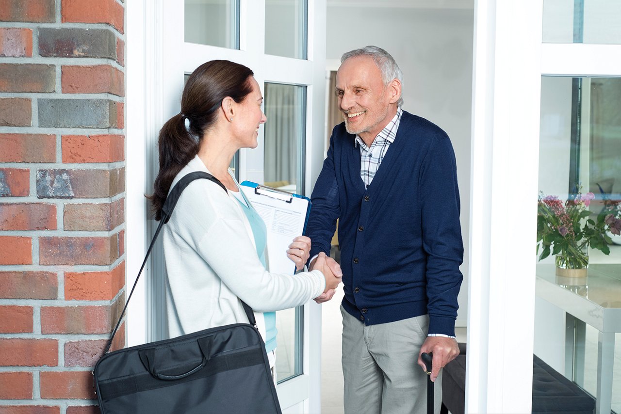 Infirmière saluant un homme âgé avec une canne à la porte d'entrée.
