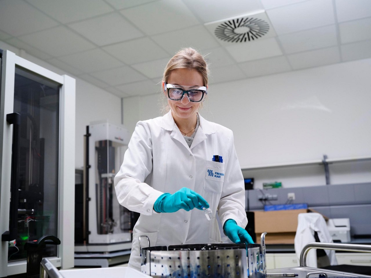 Laboratory professional wearing a white lab coat, safety goggles, and protective gloves handling a sample container in a modern laboratory environment with scientific equipment in the background.