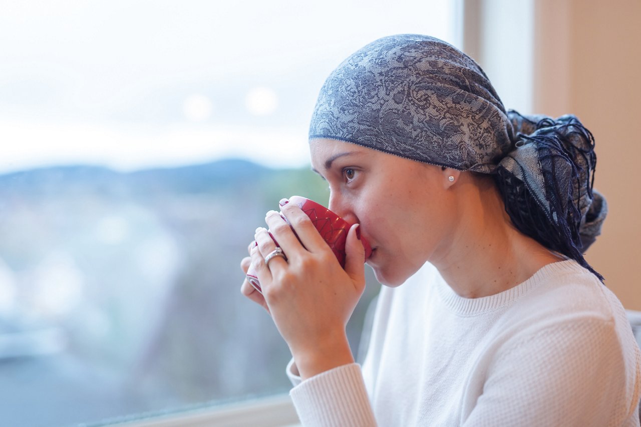 A beautiful ethnic woman in her 30s sits by her living room window with a cup of tea and looks out contemplatively. She is a cancer survivor and is wearing a headscarf.