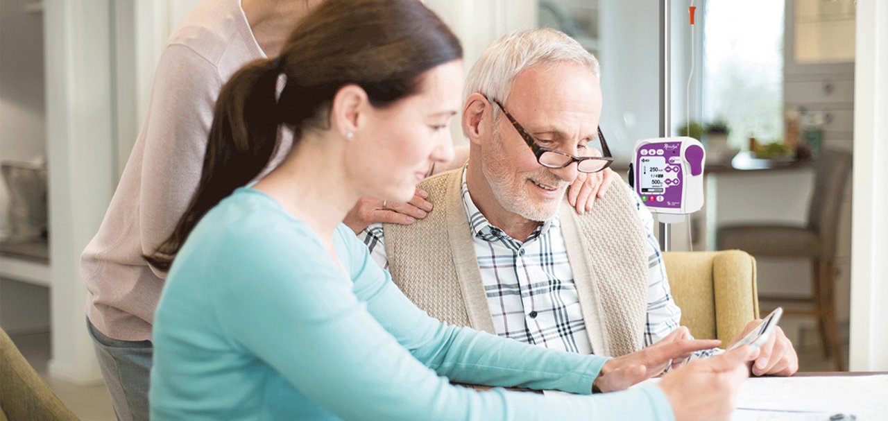 People reviewing information together in a home care setting with an Amika+ enteral feeding pump visible nearby.