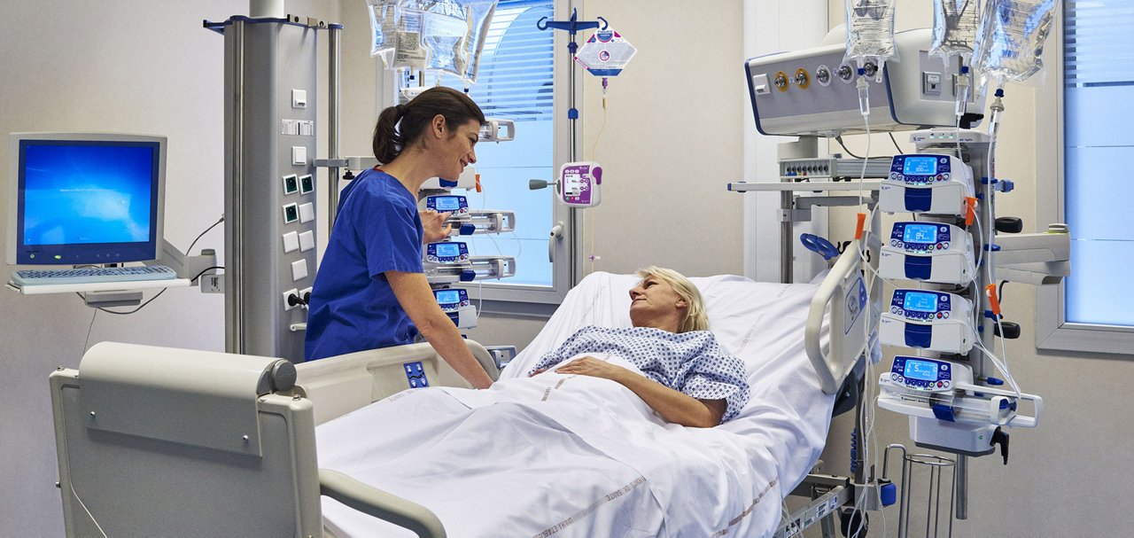 Healthcare professional caring for a patient in a hospital room equipped with multiple infusion pumps and IV therapy devices.