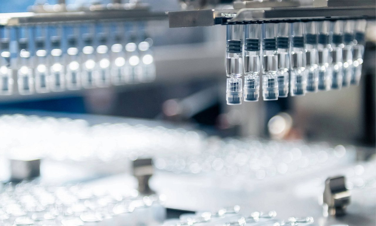 Clear glass vials arranged beneath automated pharmaceutical manufacturing equipment. Keywords