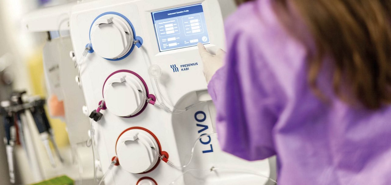 A healthcare or laboratory professional interacts with the touchscreen interface of a medical processing device. Tubing and multiple processing channels are visible.