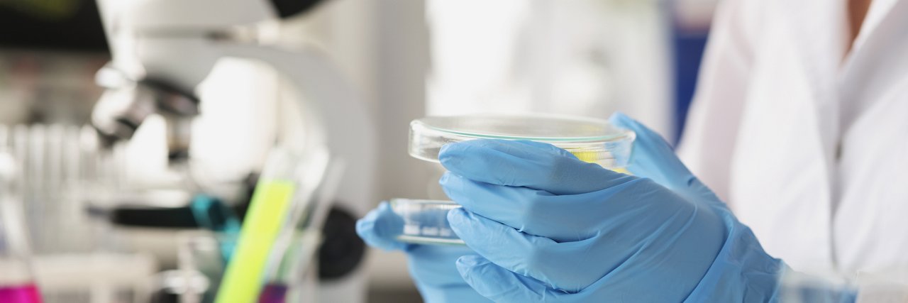 Gloved hands holding a petri dish in a lab, with a microscope and test tubes in the background