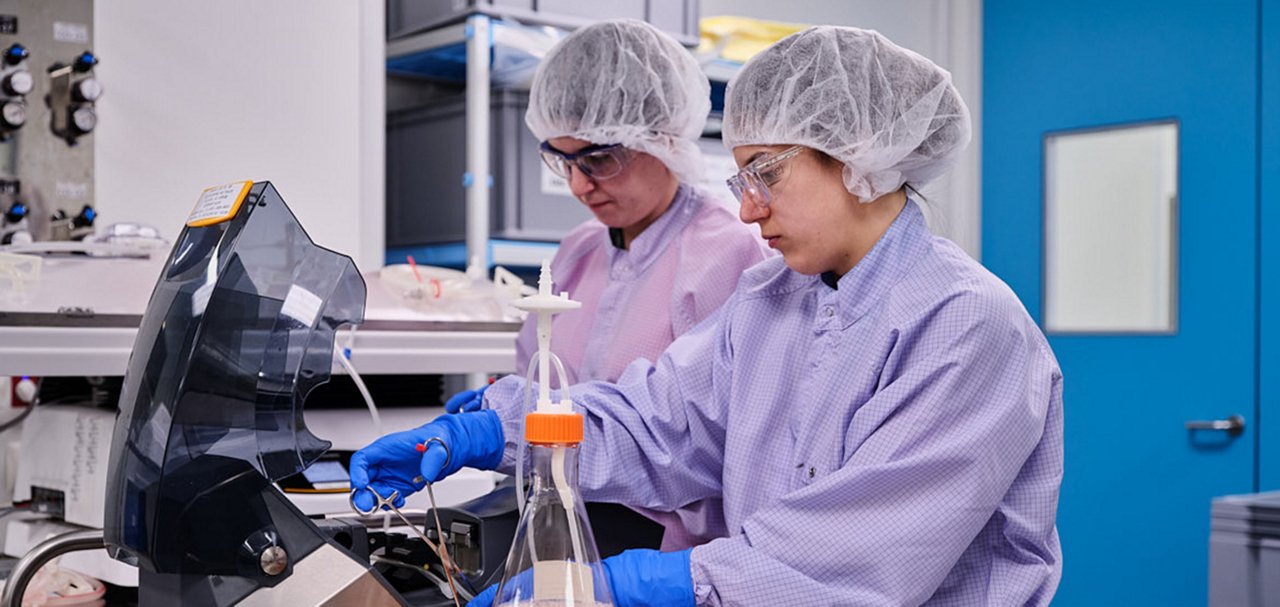 Two people in cleanroom clothing and protective eyewear working with laboratory equipment and a glass container.