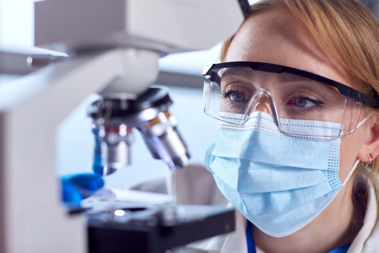 Close Up Of Female Lab Worker Wearing PPE And Safety Glasses Looking At Slide Under Microscope