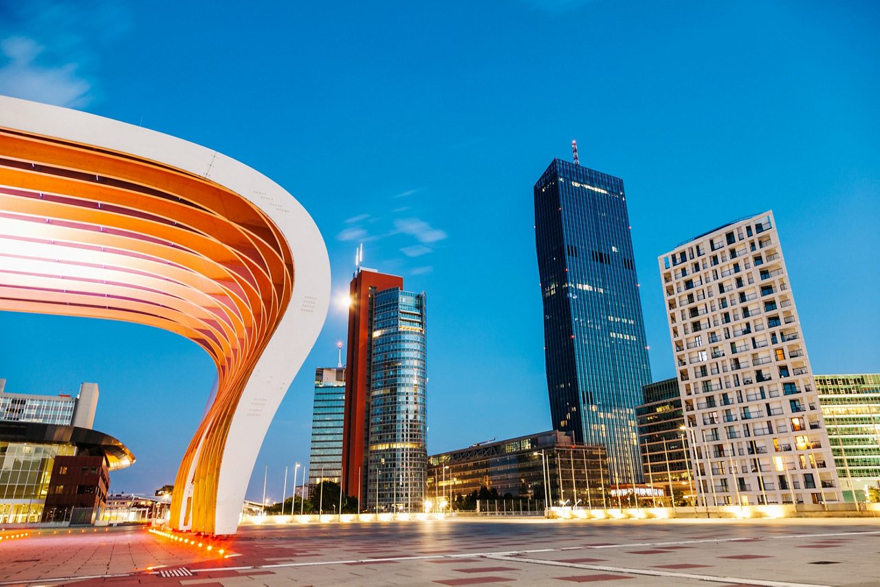 Moderne Skyline bei Dämmerung mit mehreren Hochhäusern und einer markanten, geschwungenen orange-weißen Architekturstruktur im Vordergrund unter klarem blauem Himmel.