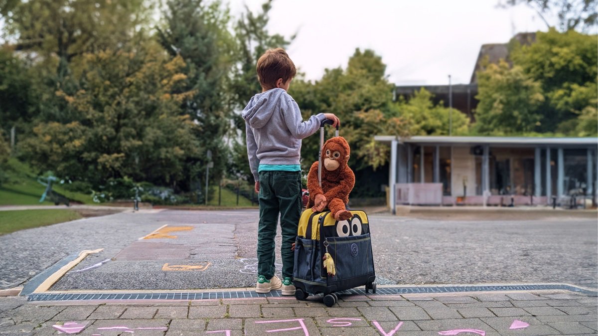 Junge steht mit dem Rücken zu uns mit seiner Tasche und einem Stoffaffen