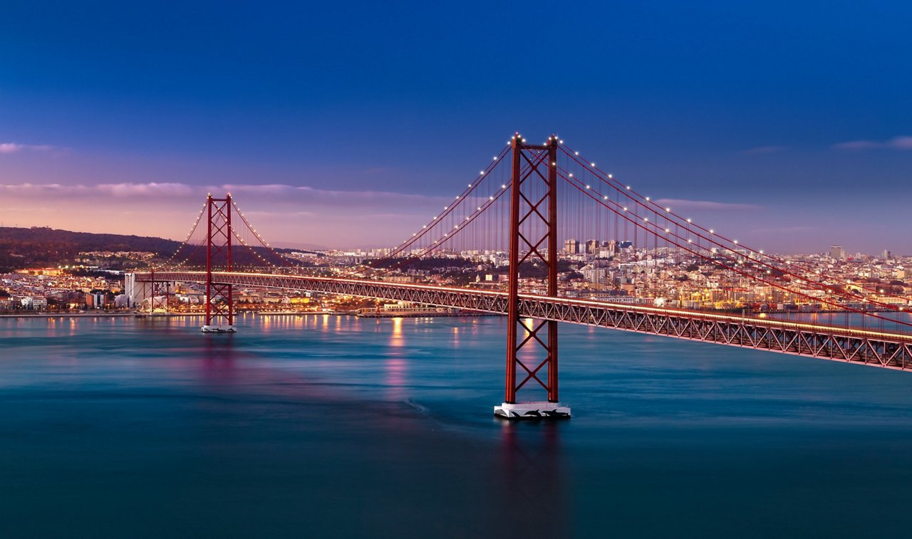Die Ponte 25 de Abril bei Abenddämmerung, beleuchtet über dem ruhigen Tejo mit Blick auf die erleuchtete Skyline von Lissabon.