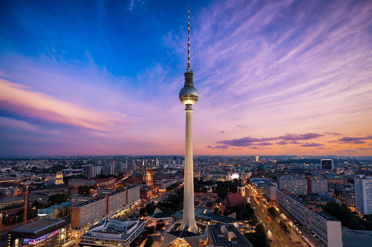 Der Berliner Fernsehturm ragt bei Sonnenuntergang über die beleuchtete Skyline von Berlin, mit Alexanderplatz und umliegenden Gebäuden unter einem farbigen Abendhimmel.