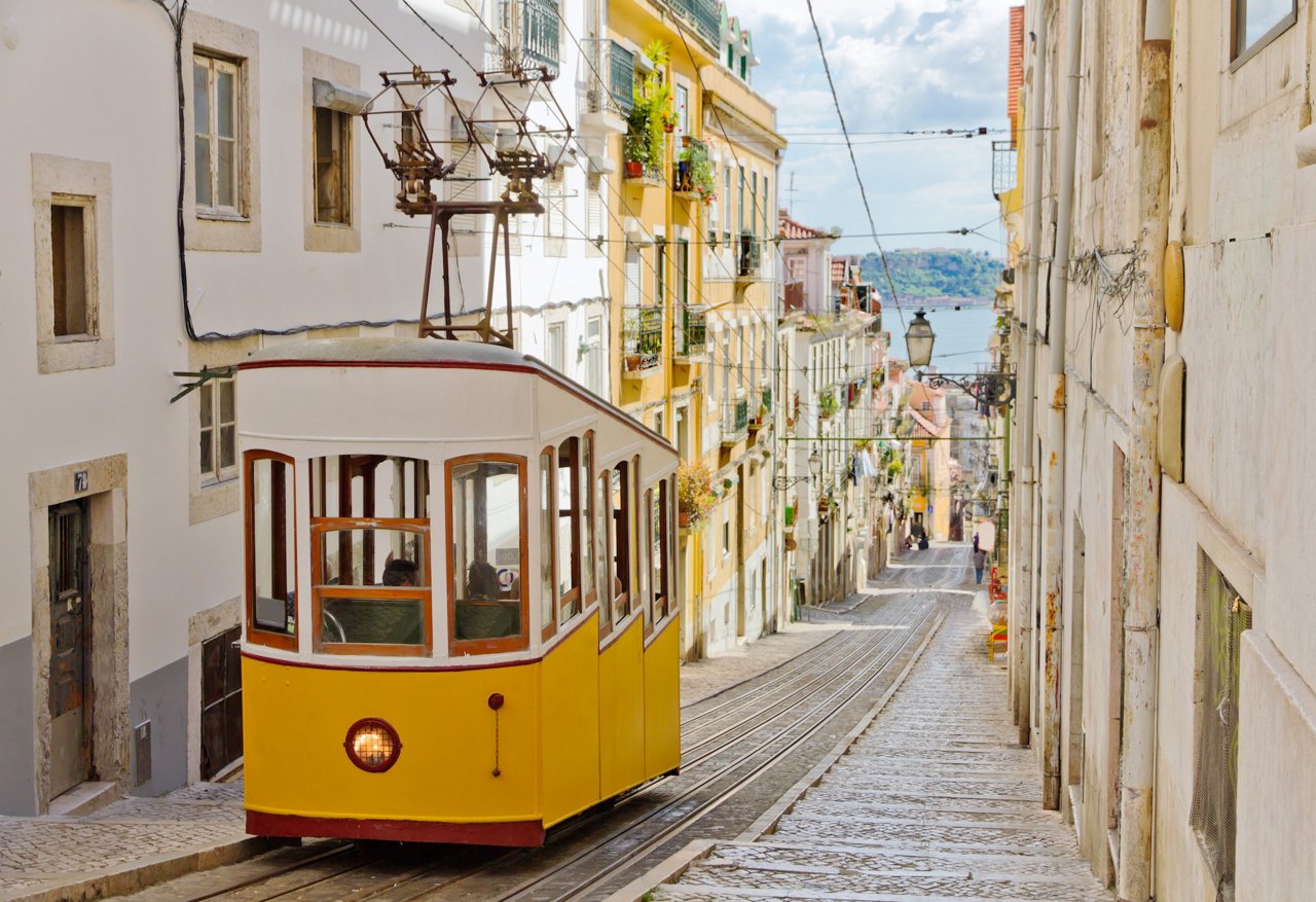 Eine traditionelle gelbe Strassenbahn fährt durch eine schmale, steile Strasse in Lissabon. Die Strasse ist von mehrstöckigen Gebäuden mit Balkonen gesäumt, im Hintergrund ist der Tejo sichtbar.