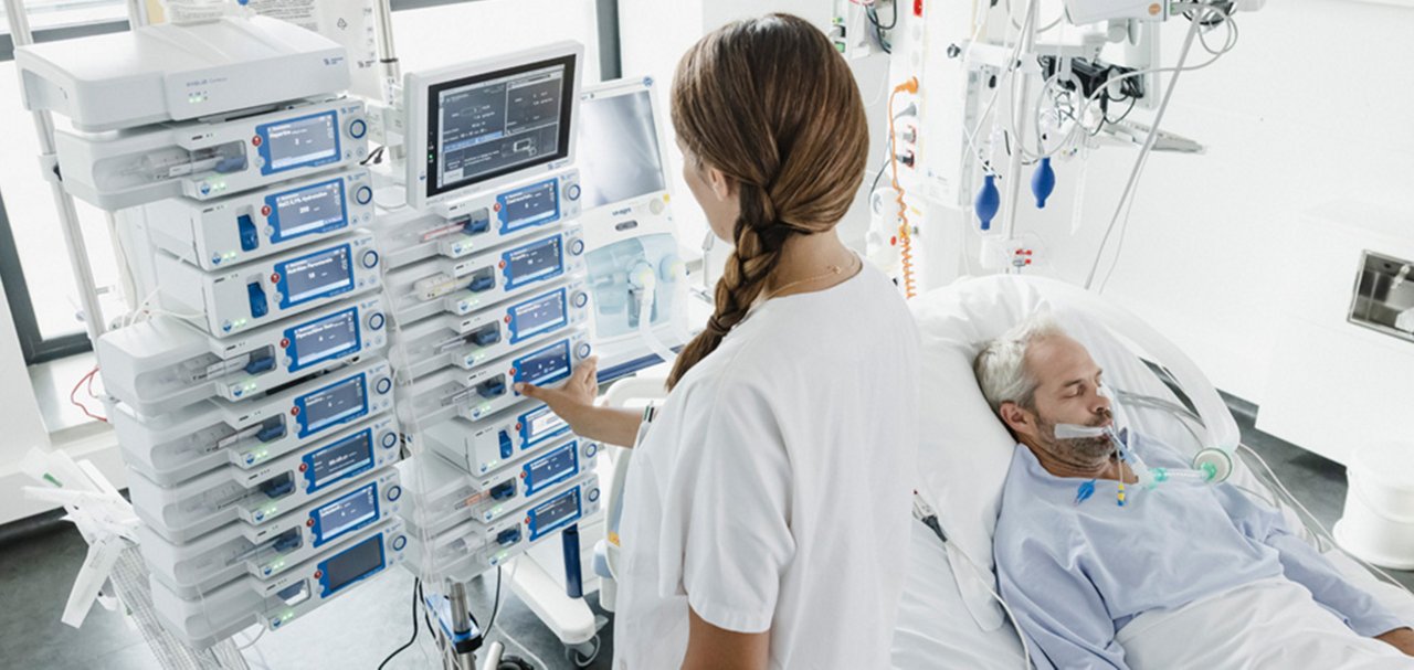Healthcare professional operating multiple infusion pumps beside a patient receiving respiratory support in a hospital care unit.
