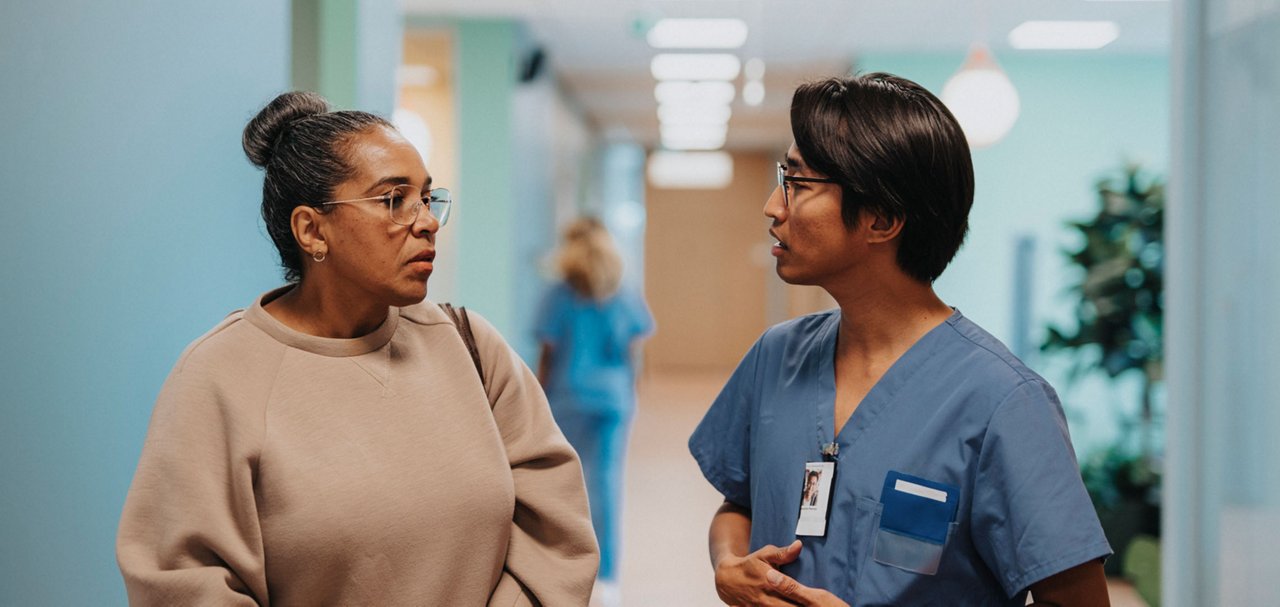 A healthcare worker in clinical attire speaks with a visitor in a healthcare corridor. Additional staff are visible in the background.