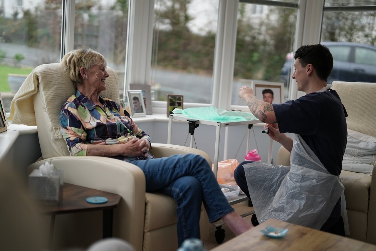 Caregiver attending to a person in a recliner chair in a bright room with large windows and framed photographs, highlighting home healthcare or personal care assistance.