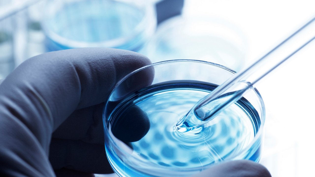 A scientist holds a petri dish uses a pipette to remove a sample of solution. In the background are test tube racks which are slightly out of focus suggesting the sterile environment of a laboratory. The hand is covered with a latex glove. A cool blue tone is the dominant color scheme