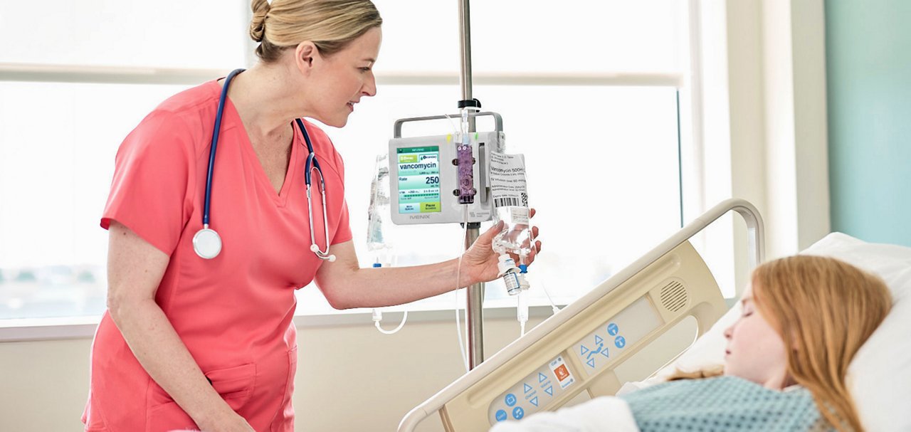 Healthcare professional adjusting an infusion pump while attending a patient in a hospital bed.