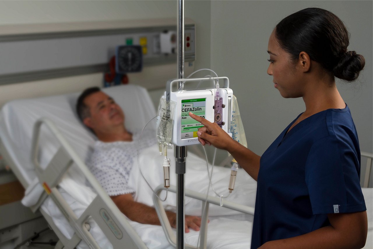 Nurse in blue scrubs adjusts IV drip for patient lying in hospital bed.
