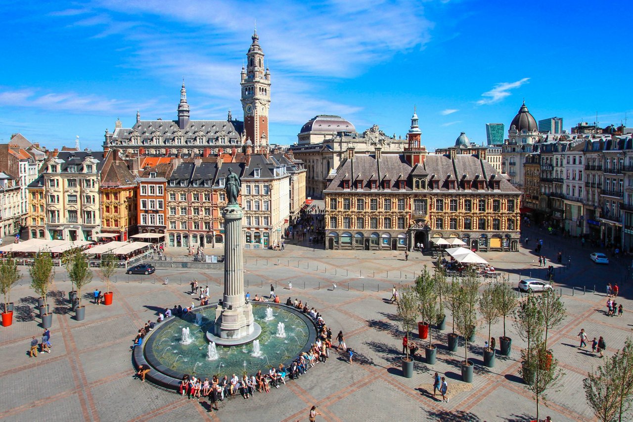 Die Grand Place in Lille mit der Colonne de la Déesse und Springbrunnen im Vordergrund, umgeben von historischen Gebäuden, der Alten Börse und dem Belfried unter einem klaren blauen Himmel.