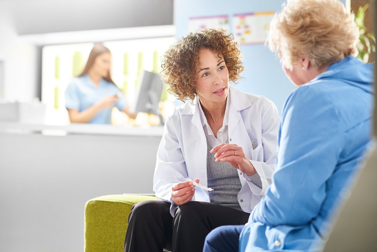 A female pharmacist sits with a senior female patient in the pharmacist consultation area and discusses her prescription and choice of medication. She is holding an a strip of the pills and chatting to the customer .In the background a female pharmacy assistant is checking orders at the counter .