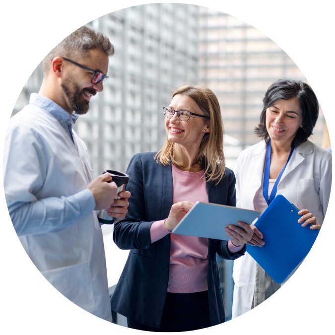 Zwei medizinische Fachkräfte in weissen Kitteln und eine Geschäftsfrau mit Tablet unterhalten sich lächelnd in einem hellen, modernen Bürogebäude mit Glasfassade.