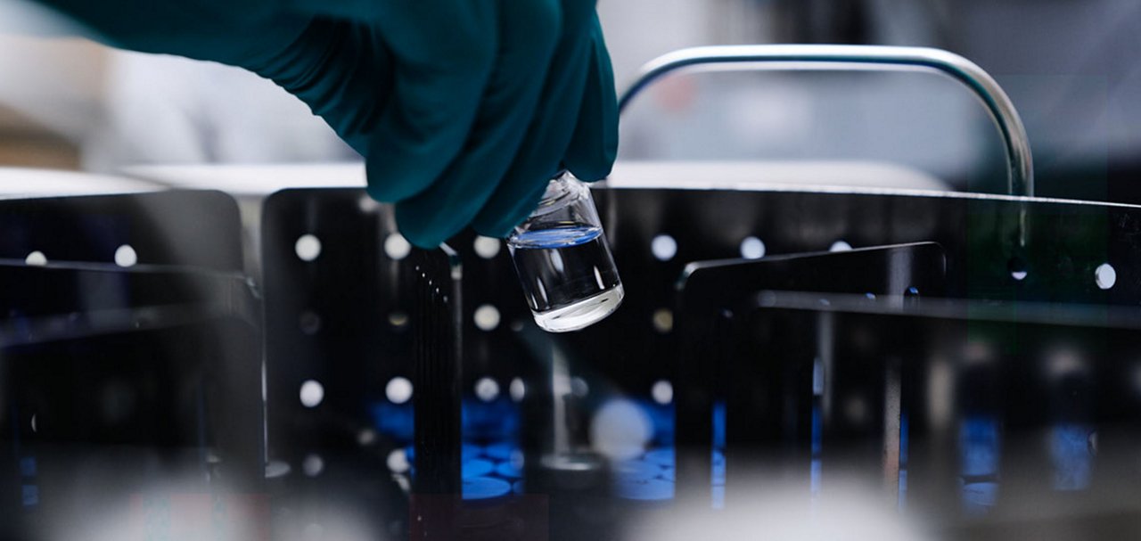 Gloved hand holding a small glass vial containing clear liquid above laboratory equipment.
