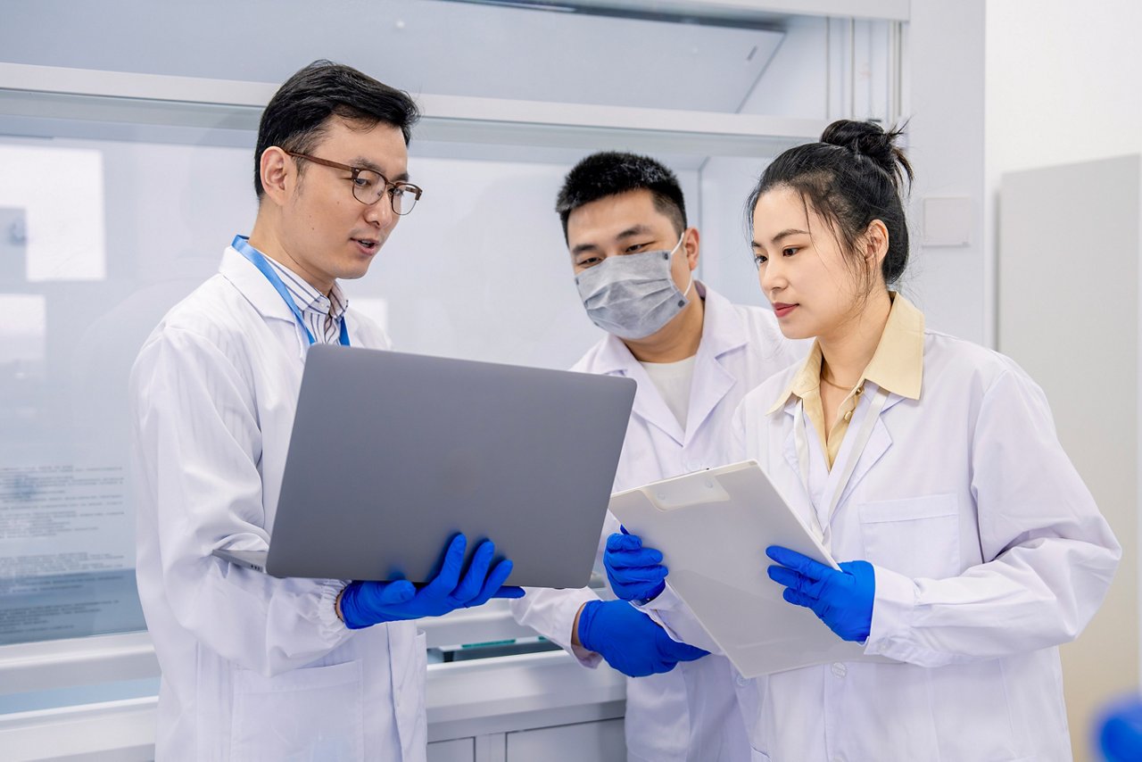 Three laboratory team members reviewing information on a laptop and clipboard.