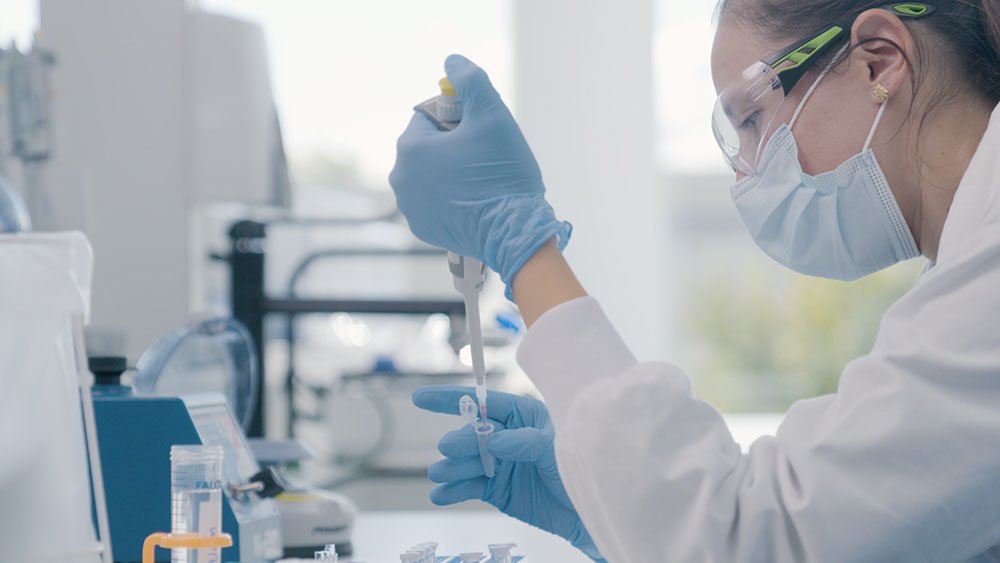 Scientist in lab coat and gloves uses pipette to transfer liquid into test tube in lab.